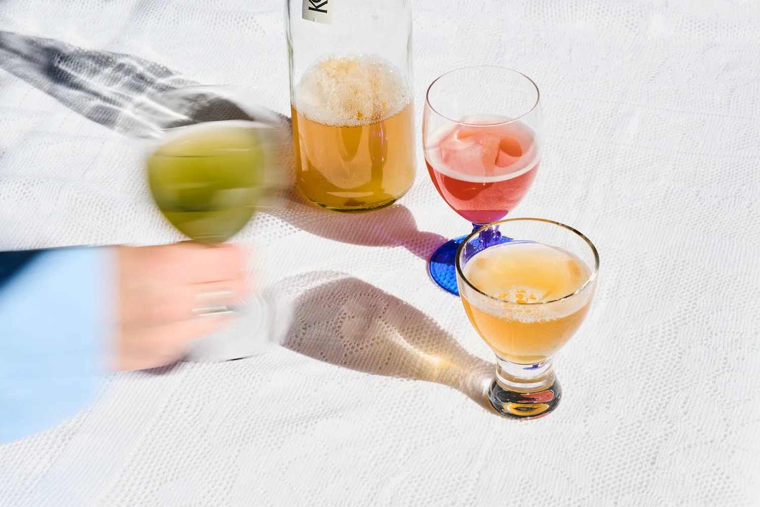 Bottle and glasses of kombucha on white linen, mid-motion