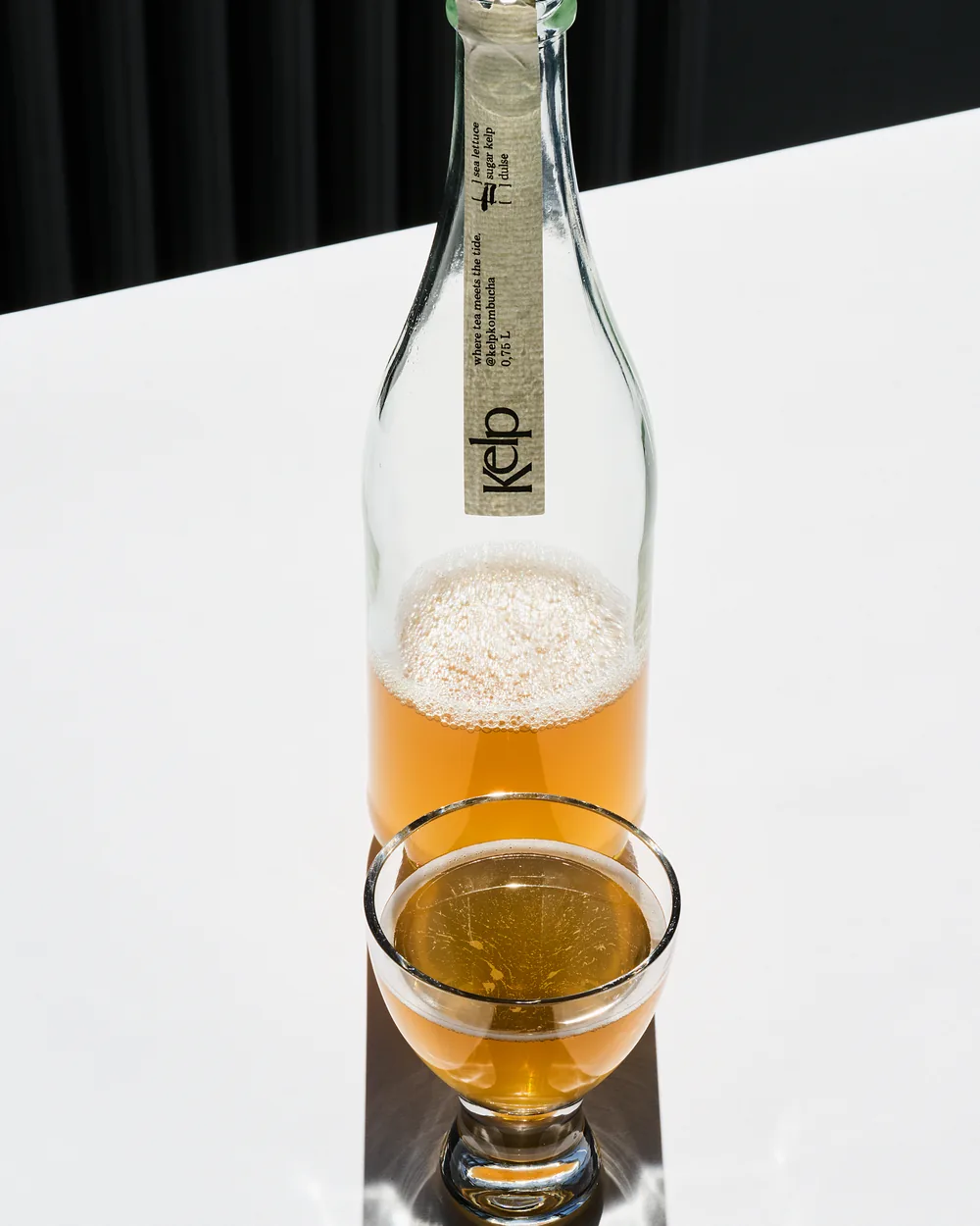 A bottle of sugar kelp kombucha with a small glass, backlit